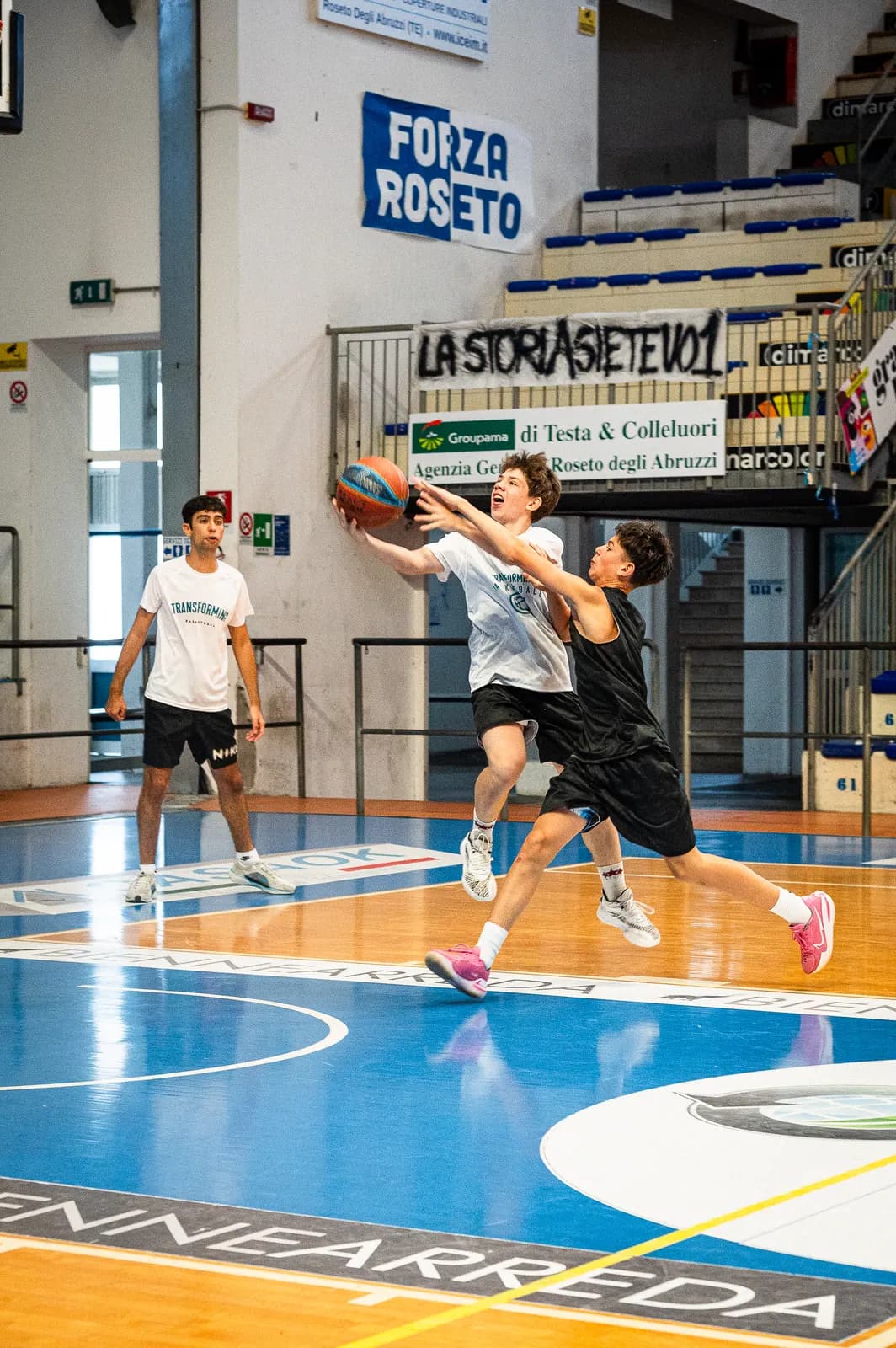 An offensive player finishing at the rim while a defender jumps to contest during a game at the 2026 Transforming Basketball camp in Roseto